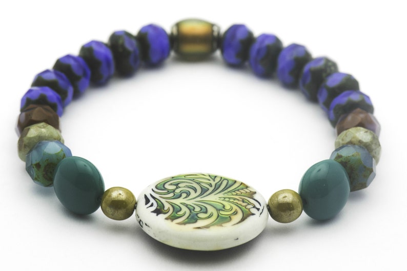 Puede incluir: Un brazalete de cuentas multicolores con un dije de dise&ntilde;o floral verde, blanco y dorado. El brazalete presenta cuentas azules, verdes, marrones y doradas.