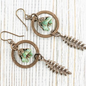 May include: A pair of handcrafted earrings featuring a bronze-coloured circular frame with a small, light green flower and a fern-like charm. The earrings have a hook closure and are displayed on a white wooden surface.