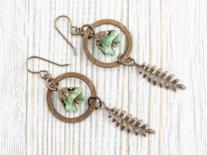 May include: A pair of handcrafted earrings featuring a bronze-coloured circular frame with a small, light green flower and a fern-like charm. The earrings have a hook closure and are displayed on a white wooden surface.