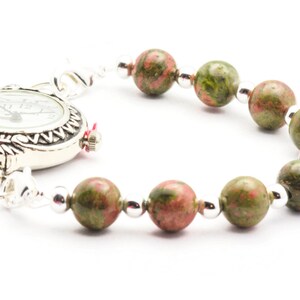May include: A silver watch with a white face and black Roman numeral markings is attached to a bracelet made of green and pink speckled beads.