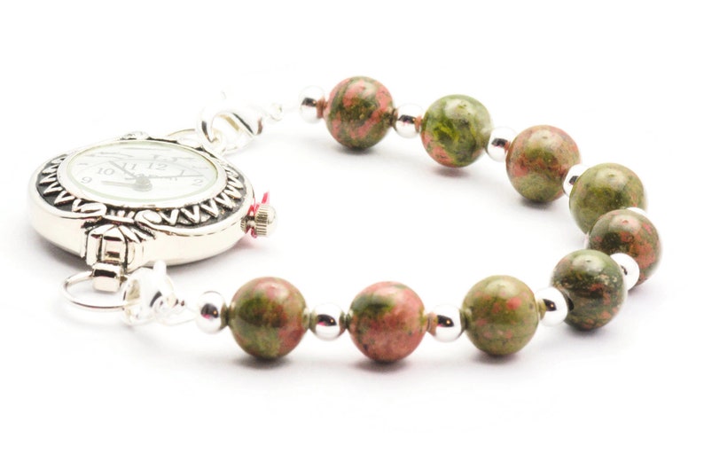 May include: A silver watch with a white face and black Roman numeral markings is attached to a bracelet made of green and pink speckled beads.