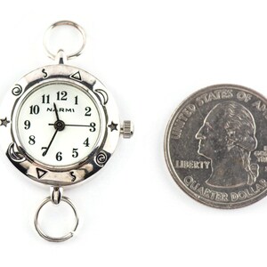 May include: A silver-toned analog watch with a white face, black numerals, and the brand name Narmi. The bezel features engraved celestial symbols including stars, moons, and spirals. A silver ring is attached to the top and bottom. A U.S. quarter is shown for scale.