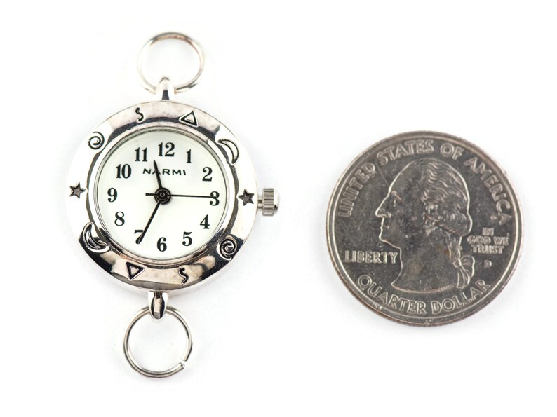 May include: A silver-toned analog watch with a white face, black numerals, and the brand name Narmi. The bezel features engraved celestial symbols including stars, moons, and spirals. A silver ring is attached to the top and bottom. A U.S. quarter is shown for scale.