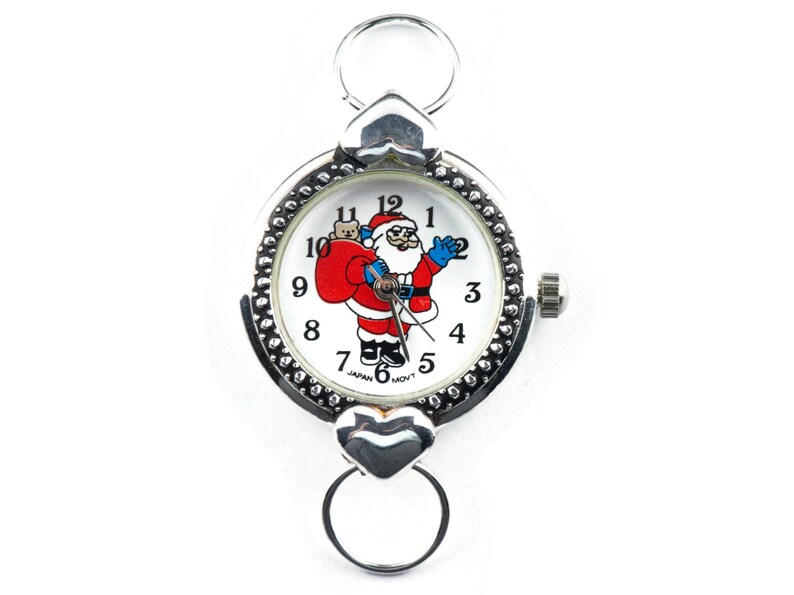 Peut inclure: Une montre argent&eacute;e avec un cadran blanc repr&eacute;sentant un P&egrave;re No&euml;l en dessin anim&eacute; tenant un sac de cadeaux. La montre a un fermoir en forme de c&oelig;ur et une cha&icirc;ne argent&eacute;e.