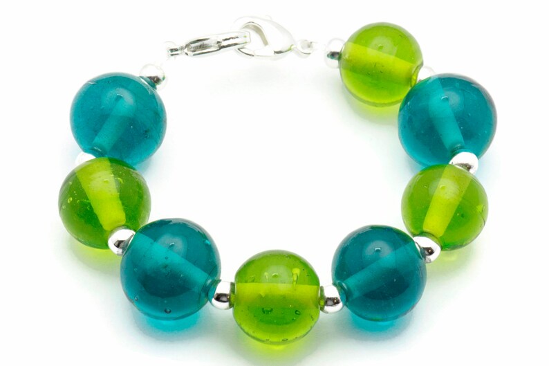 Puede incluir: Un brazalete hecho de cuentas de vidrio alternas de color verde azulado y verde con separadores plateados y un cierre plateado.