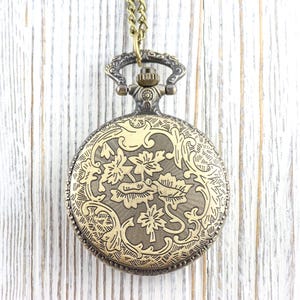 May include: An antique brass pocket watch with an elaborate floral pattern on the front. It hangs from a matching chain. The background is a light-coloured, weathered wooden surface.