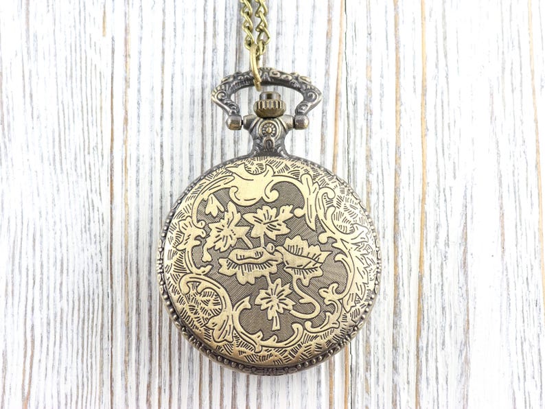 May include: An antique brass pocket watch with an elaborate floral pattern on the front. It hangs from a matching chain. The background is a light-coloured, weathered wooden surface.