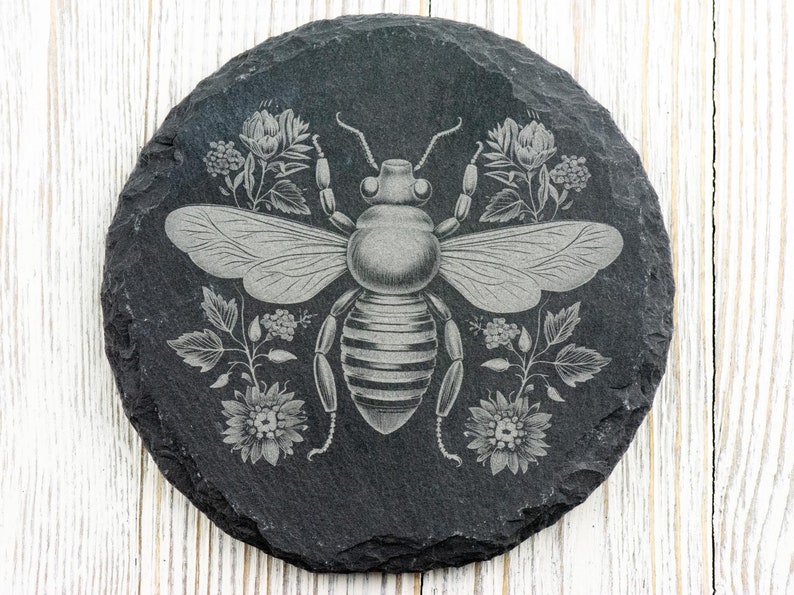 Op de afbeelding: Een zwarte leisteen onderzetter met een lasergegraveerd ontwerp van een bij omringd door bloemen. De bij is grijs en heeft een gestreepte buik. De bloemen zijn wit en grijs.
