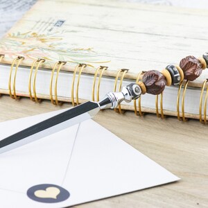 Op de afbeelding: Een zilveren briefopener met een bruin en zwart geperld handvat. Het handvat is gemaakt van hout en heeft een bladontwerp. De briefopener ligt op een witte envelop met een zwarte hartensticker.