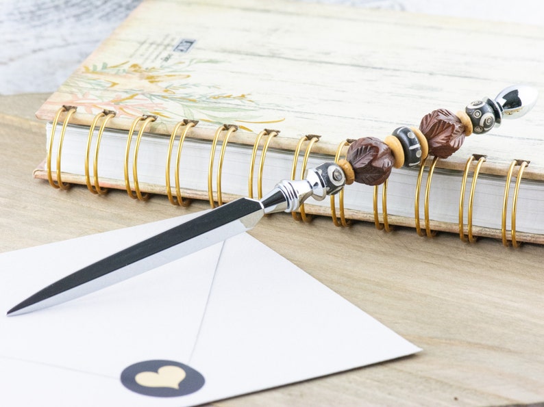 Op de afbeelding: Een zilveren briefopener met een bruin en zwart geperld handvat. Het handvat is gemaakt van hout en heeft een bladontwerp. De briefopener ligt op een witte envelop met een zwarte hartensticker.