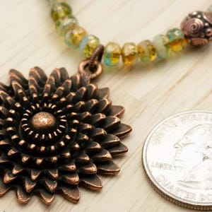 Pu&ograve; includere: Una collana con pendente color rame con un grande motivo floreale dettagliato. La collana presenta un filo di perline con perline verdi e gialle. Una moneta &egrave; accanto al pendente per il confronto delle dimensioni.