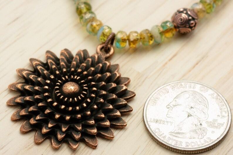 Pu&ograve; includere: Una collana con pendente color rame con un grande motivo floreale dettagliato. La collana presenta un filo di perline con perline verdi e gialle. Una moneta &egrave; accanto al pendente per il confronto delle dimensioni.