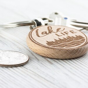K&ouml;nnte beinhalten: Ein Holzschl&uuml;sselanh&auml;nger mit dem eingravierten Schriftzug "Lake Life" sowie einem Wald- und Wassermotiv. Der Schl&uuml;sselanh&auml;nger ist rund und an einem silbernen Schl&uuml;sselring befestigt. Eine M&uuml;nze liegt neben dem Schl&uuml;sselanh&auml;nger.