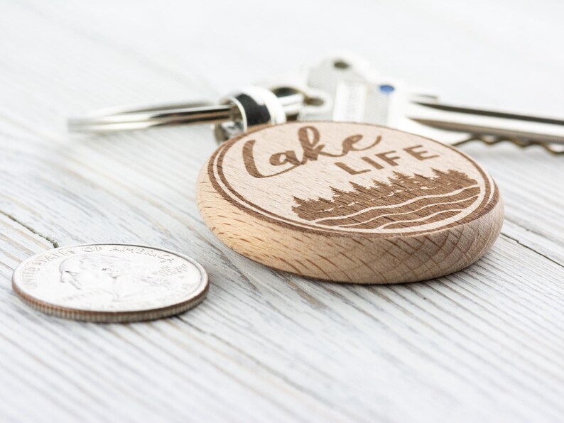 K&ouml;nnte beinhalten: Ein Holzschl&uuml;sselanh&auml;nger mit dem eingravierten Schriftzug "Lake Life" sowie einem Wald- und Wassermotiv. Der Schl&uuml;sselanh&auml;nger ist rund und an einem silbernen Schl&uuml;sselring befestigt. Eine M&uuml;nze liegt neben dem Schl&uuml;sselanh&auml;nger.