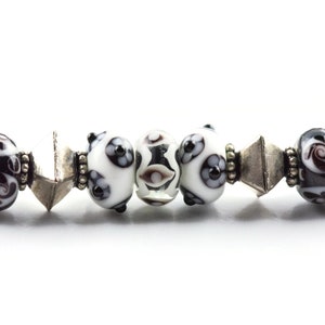 May include: A close-up of a black and white beaded bracelet with silver accents. The bracelet features a variety of round beads with intricate patterns.