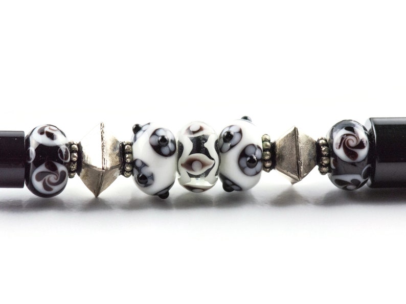 May include: A close-up of a black and white beaded bracelet with silver accents. The bracelet features a variety of round beads with intricate patterns.