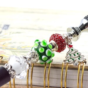May include: A decorative pen with a black barrel and a silver-colored top, adorned with various beads and charms. The charms include a green and white bead, a red beaded sphere, and a Christmas tree charm. The pen is resting on a notebook.