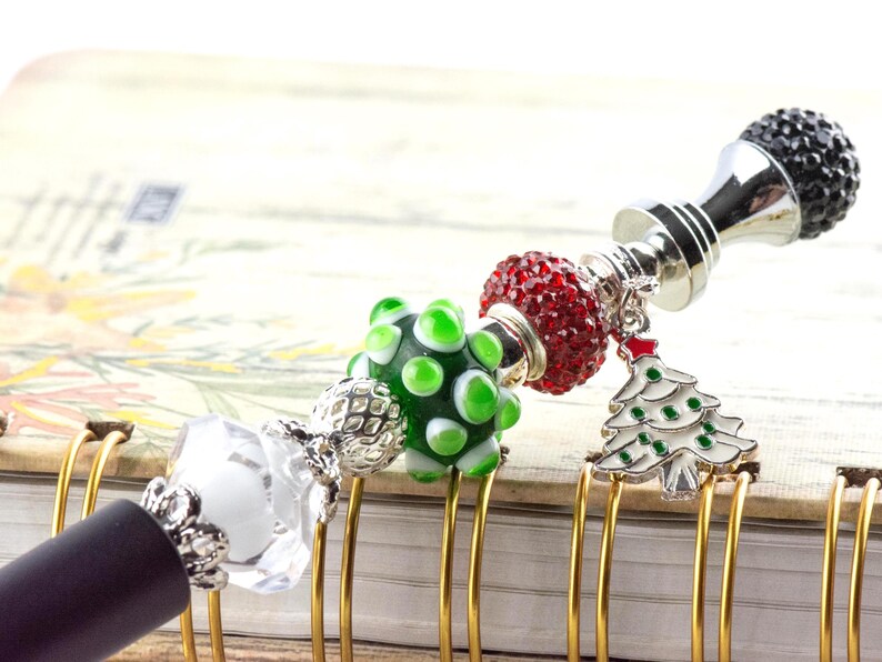 May include: A decorative pen with a black barrel and a silver-colored top, adorned with various beads and charms. The charms include a green and white bead, a red beaded sphere, and a Christmas tree charm. The pen is resting on a notebook.