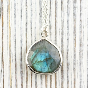 Pu&ograve; includere: Una collana con pendente in argento con una pietra preziosa di labradorite sfaccettata a forma di goccia. La pietra mostra sfumature iridescenti blu e verdi. Il pendente &egrave; appeso a una delicata catena d'argento, su uno sfondo di legno bianco.