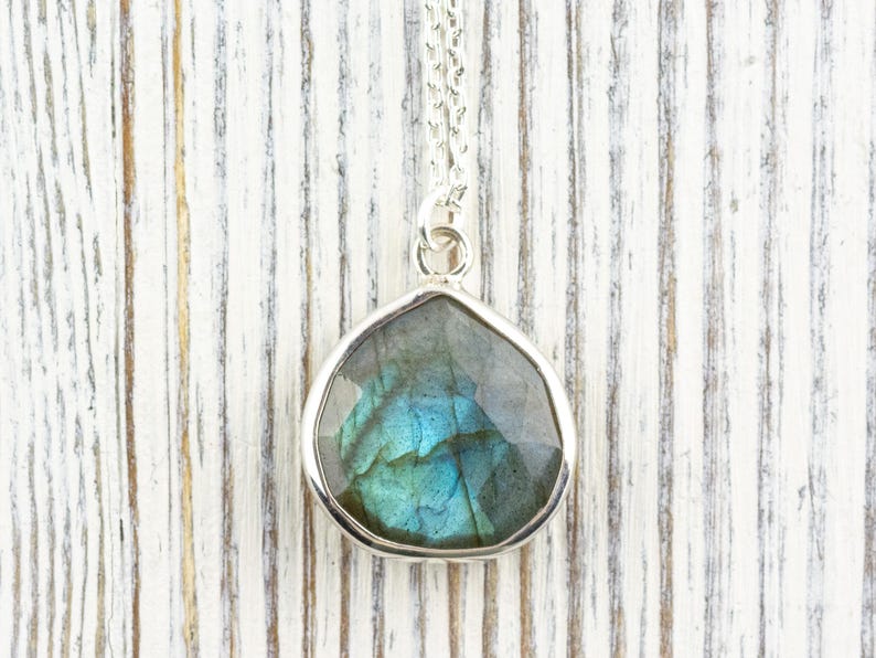 Pu&ograve; includere: Una collana con pendente in argento con una pietra preziosa di labradorite sfaccettata a forma di goccia. La pietra mostra sfumature iridescenti blu e verdi. Il pendente &egrave; appeso a una delicata catena d'argento, su uno sfondo di legno bianco.