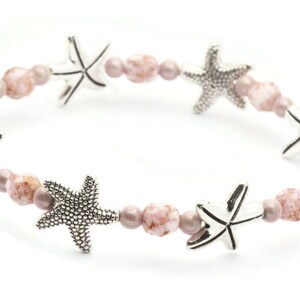 K&ouml;nnte beinhalten: Dehnbare Armband mit silbernen Seestern-Anh&auml;ngern und einer Mischung aus rosa und mauvefarbenen Perlen. Das Armband hat ein maritimes Thema und ist ein sch&ouml;nes Accessoire f&uuml;r den Strand oder den Alltag.
