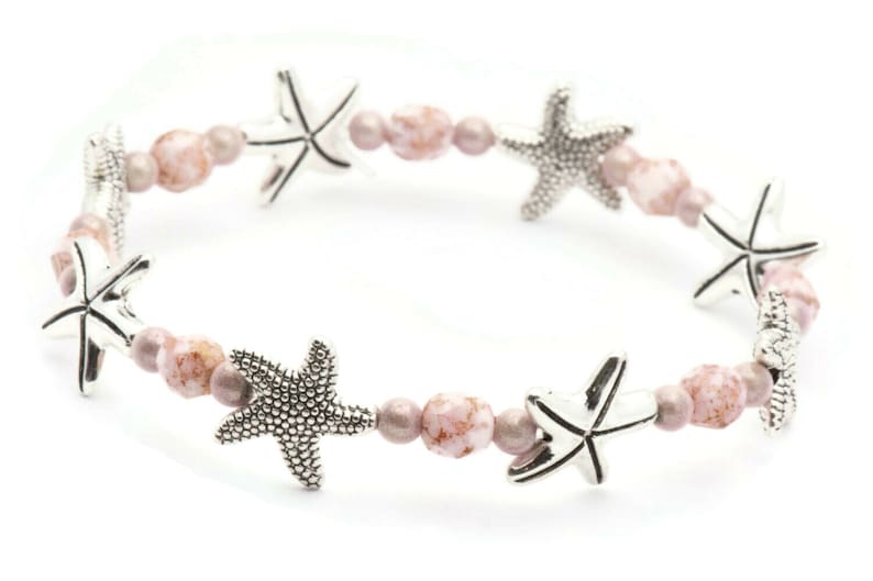 K&ouml;nnte beinhalten: Dehnbare Armband mit silbernen Seestern-Anh&auml;ngern und einer Mischung aus rosa und mauvefarbenen Perlen. Das Armband hat ein maritimes Thema und ist ein sch&ouml;nes Accessoire f&uuml;r den Strand oder den Alltag.