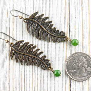 K&ouml;nnte beinhalten: Ein Paar bronzefarbene Ohrringe in Form von Farnbl&auml;ttern mit gr&uuml;nen Edelstein-Akzenten. Die Ohrringe sind etwa so gro&szlig; wie eine US-amerikanische 25-Cent-M&uuml;nze.