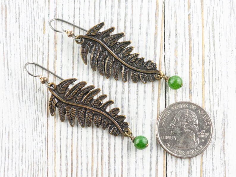 K&ouml;nnte beinhalten: Ein Paar bronzefarbene Ohrringe in Form von Farnbl&auml;ttern mit gr&uuml;nen Edelstein-Akzenten. Die Ohrringe sind etwa so gro&szlig; wie eine US-amerikanische 25-Cent-M&uuml;nze.