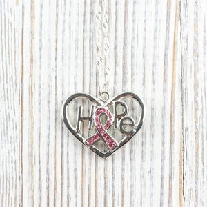 Pode incluir: Colar prateado com um pingente em forma de cora&ccedil;&atilde;o. O pingente apresenta a palavra "HOPE" e uma fita rosa. O colar est&aacute; sobre um fundo de madeira branca.