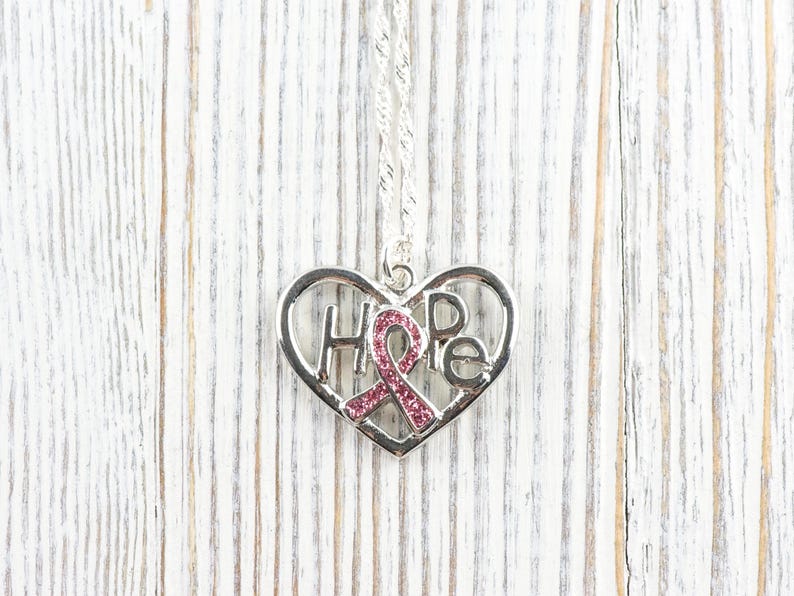 Pode incluir: Colar prateado com um pingente em forma de cora&ccedil;&atilde;o. O pingente apresenta a palavra "HOPE" e uma fita rosa. O colar est&aacute; sobre um fundo de madeira branca.