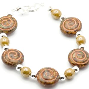 May include: A bracelet with brown and orange spiral-patterned beads, olive-green beads, and small silver beads. The bracelet is connected with a silver clasp. The jewellery is shown against a white backdrop.