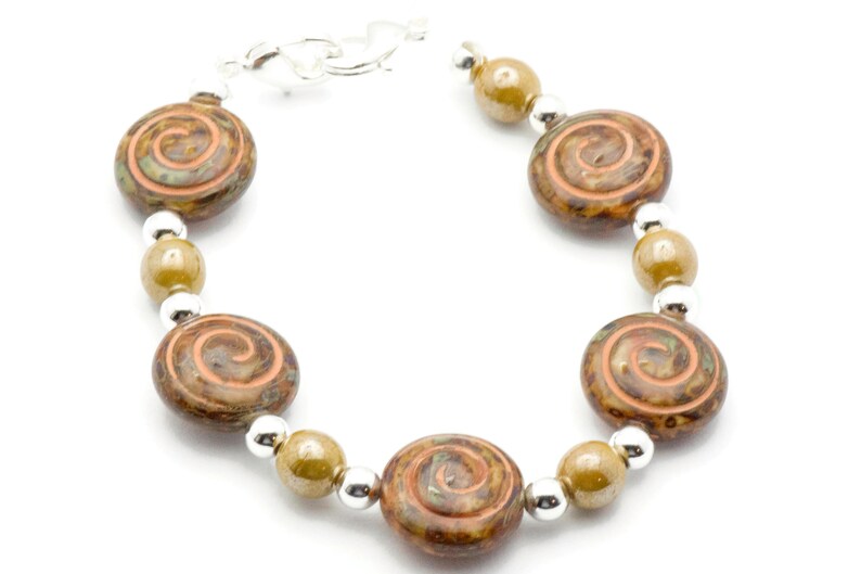 May include: A bracelet with brown and orange spiral-patterned beads, olive-green beads, and small silver beads. The bracelet is connected with a silver clasp. The jewellery is shown against a white backdrop.