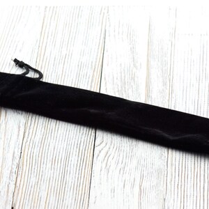 May include: A black velvet pen pouch with a drawstring closure. The pouch is long and slender, designed to hold a single pen or pencil. The background is a light-colored, textured wooden surface.