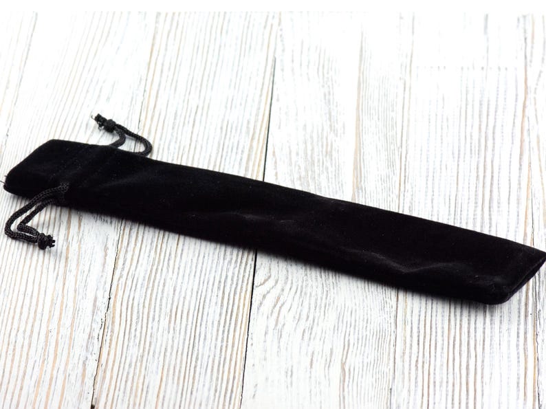 May include: A black velvet pen pouch with a drawstring closure. The pouch is long and slender, designed to hold a single pen or pencil. The background is a light-colored, textured wooden surface.
