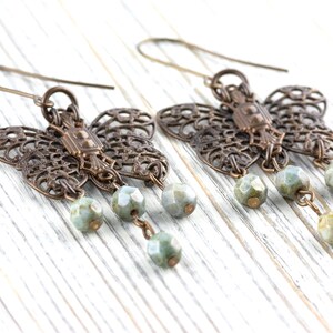 May include: A pair of bronze-colored butterfly earrings with intricate filigree wings. Each earring features three dangling, faceted, pale green beads. The earrings are displayed on a white wooden surface.