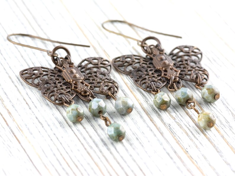 May include: A pair of bronze-colored butterfly earrings with intricate filigree wings. Each earring features three dangling, faceted, pale green beads. The earrings are displayed on a white wooden surface.