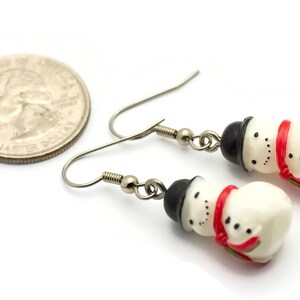 May include: A pair of silver dangle earrings featuring a snowman charm with a black hat, white body, red scarf, and black eyes. The earrings are shown next to a US quarter for size reference.
