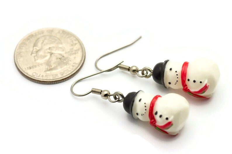May include: A pair of silver dangle earrings featuring a snowman charm with a black hat, white body, red scarf, and black eyes. The earrings are shown next to a US quarter for size reference.