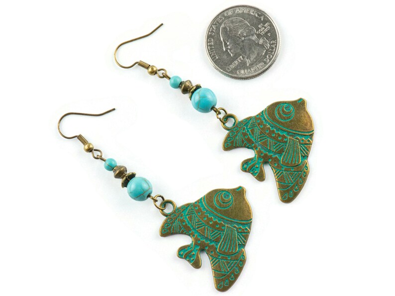 K&ouml;nnte beinhalten: Ein Paar t&uuml;rkisfarbene und bronzene Fische-Ohrringe mit filigranen Details. Jeder Ohrring hat eine t&uuml;rkisfarbene Perle und einen bronzenen Fisch-Anh&auml;nger mit t&uuml;rkisfarbener Emaille-Oberfl&auml;che.