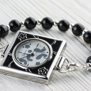 May include: A square-shaped watch with a black face and silver trim. The watch is attached to a black beaded bracelet.