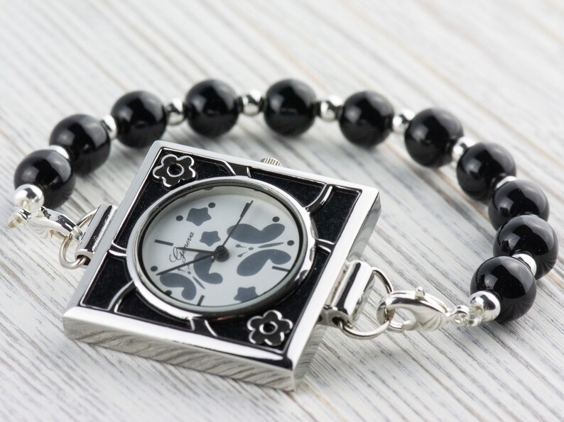 May include: A square-shaped watch with a black face and silver trim. The watch is attached to a black beaded bracelet.