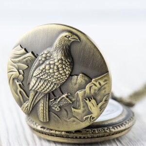 May include: A gold-toned pocket watch with a detailed engraving of a bird on the front. The watch is open, revealing a white clock face.