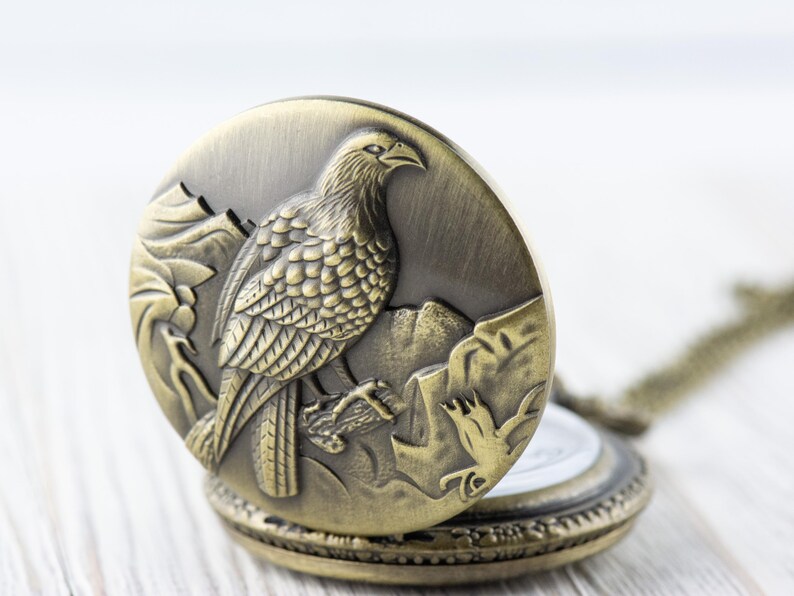 May include: A gold-toned pocket watch with a detailed engraving of a bird on the front. The watch is open, revealing a white clock face.