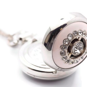 May include: A silver pocket watch with a pink face and a circle of rhinestones around a larger rhinestone in the center.