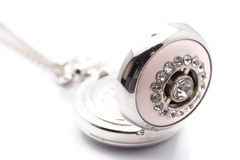 May include: A silver pocket watch with a pink face and a circle of rhinestones around a larger rhinestone in the center.