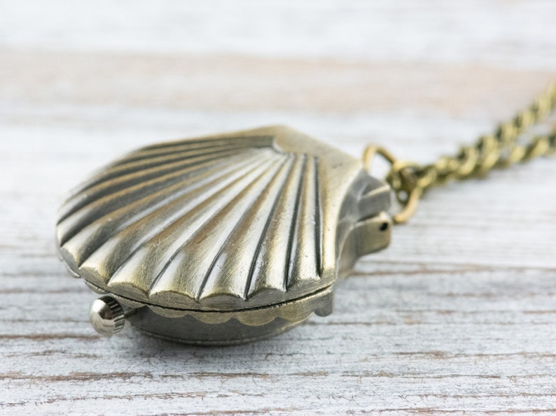 May include: A silver-toned seashell pendant necklace with a chain. The pendant is shaped like a seashell with a detailed design.