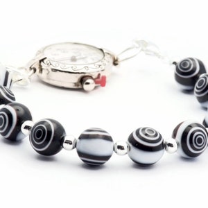 K&ouml;nnte beinhalten: Eine silberne Uhr mit wei&szlig;em Zifferblatt und schwarzen Zeigern, die an einem Armband mit schwarz-wei&szlig;en Perlen befestigt ist.
