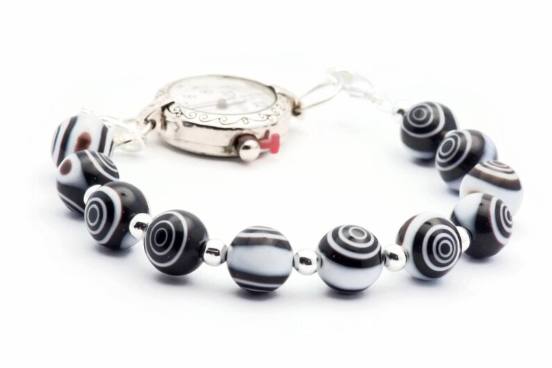 K&ouml;nnte beinhalten: Eine silberne Uhr mit wei&szlig;em Zifferblatt und schwarzen Zeigern, die an einem Armband mit schwarz-wei&szlig;en Perlen befestigt ist.