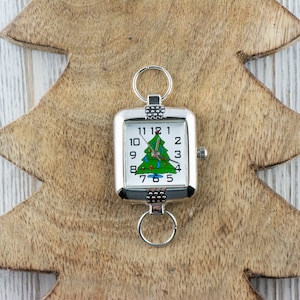 Puede incluir: Un reloj cuadrado plateado con un dise&ntilde;o de &aacute;rbol de Navidad en la esfera, con un &aacute;rbol verde con adornos rojos. El reloj est&aacute; sujeto a un llavero y se encuentra sobre un fondo de madera con forma de &aacute;rbol de Navidad.