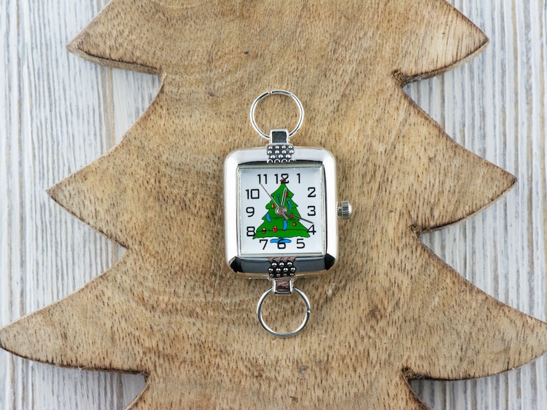 Puede incluir: Un reloj cuadrado plateado con un dise&ntilde;o de &aacute;rbol de Navidad en la esfera, con un &aacute;rbol verde con adornos rojos. El reloj est&aacute; sujeto a un llavero y se encuentra sobre un fondo de madera con forma de &aacute;rbol de Navidad.